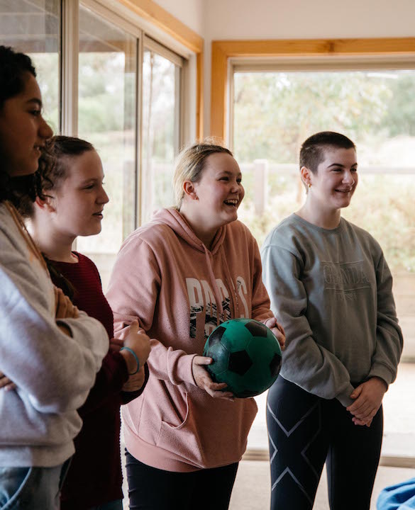 Four Young Carers stand together as part of a circle. One holds a blue soccer ball to her chest and she is laughing and looking off towards the right. The others stand, hands by their side or crossed together, looking off in the same direction and smiling. They are in a room with windows looking out to lots of trees and a campsite.